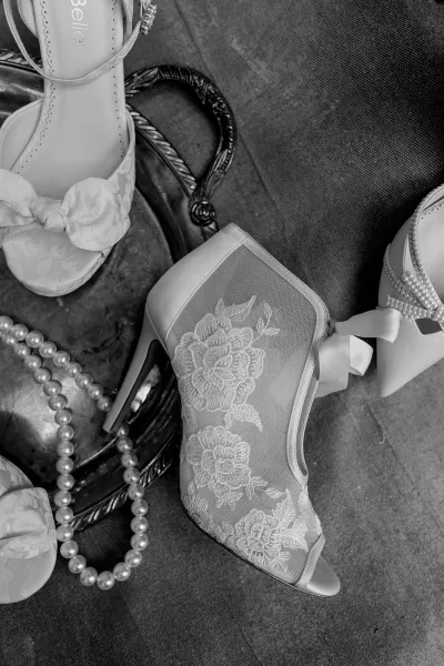 Wedding shoes, lace bridal booties with pearl accents arranged on a metal tray over a fabric surface.