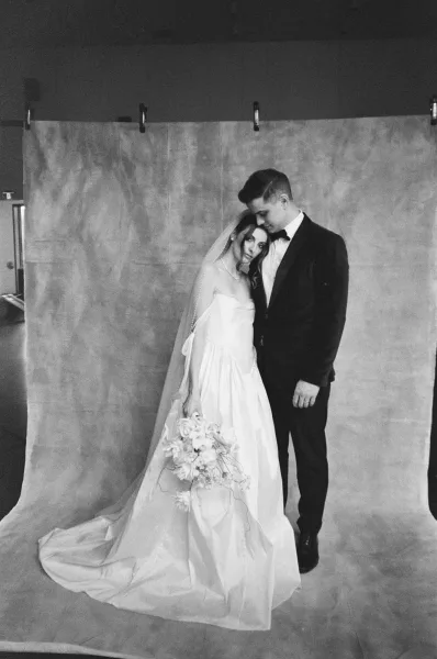 Couple portrait, a black and white wedding portrait with the pair touching foreheads, her veil over shoulder, against a studio canvas backdrop.