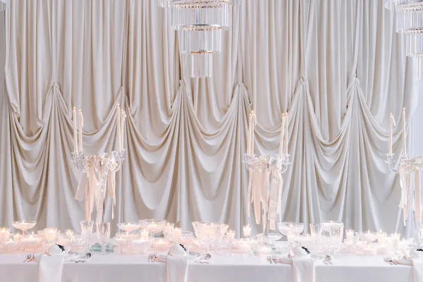 Wedding tablescape on an elegant white reception table with crystal glassware and a draped ivory curtain backdrop.