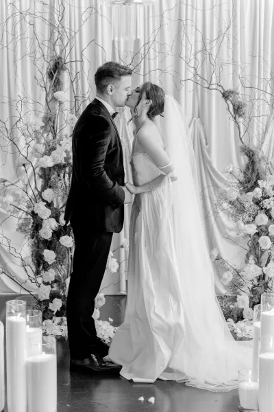 Wedding kiss portrait of the bride and groom sharing their ceremony first kiss beneath a draped fabric backdrop, candles flickering around them.