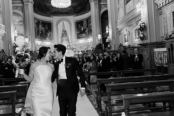 Wedding recessional as bride and groom walk down the church aisle, bouquet and veil flowing, guests cheering beneath chandeliers