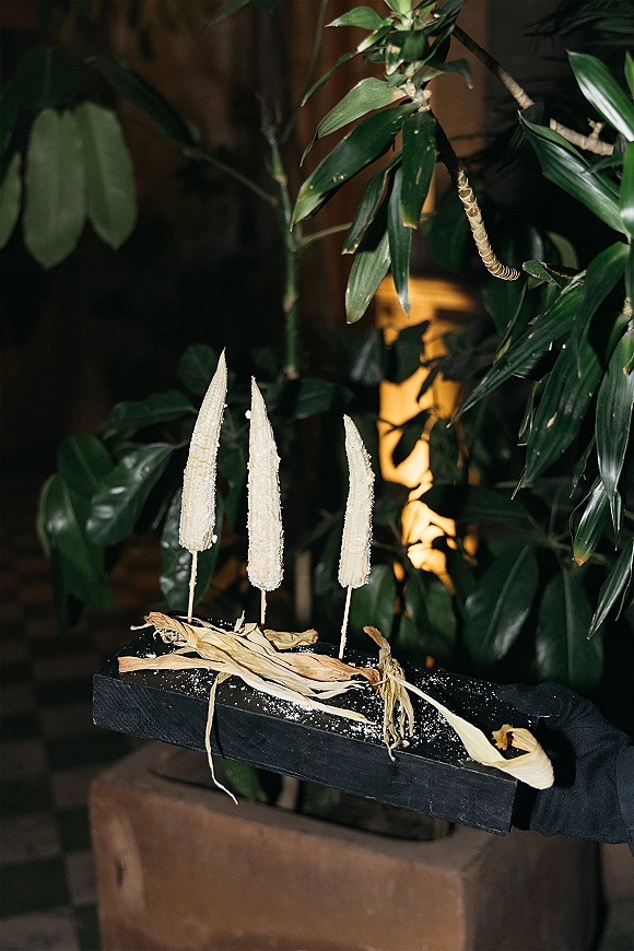 Wedding dessert display of corn cob treats dusted with powdered sugar on a black tray, styled with dried husks under warm indoor light