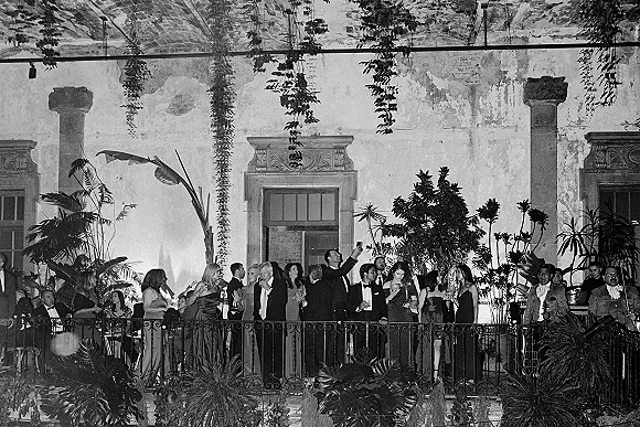 Wedding reception guests cocktail hour in tuxedos and evening gowns raise champagne glasses on a villa balcony terrace by tall windows