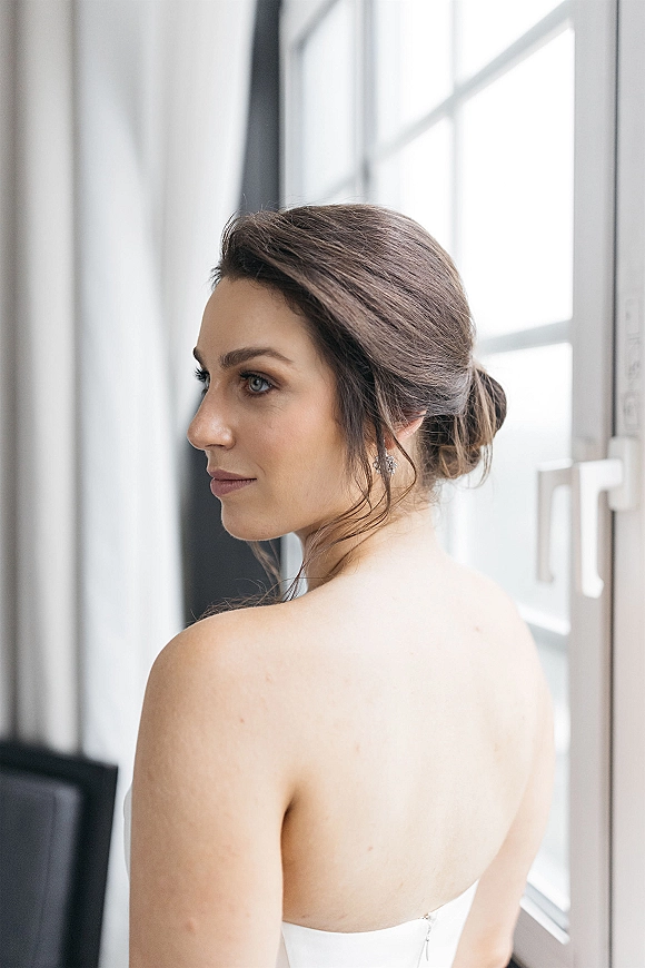 Bridal portrait of bride looking over shoulder in a strapless wedding dress with stud earrings, softly lit by a large window indoors