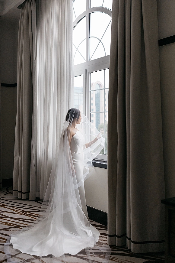 Bridal portrait of a bride by window in a strapless satin wedding dress, veil over her face and long train in a hotel room with drapes
