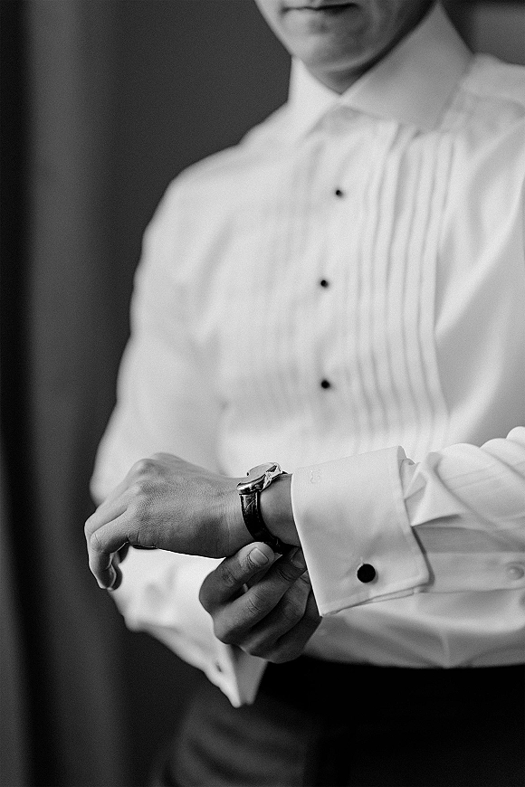 Groom getting ready, adjusting watch over a pleated tuxedo shirt with black stud buttons and French cuffs against a plain wall