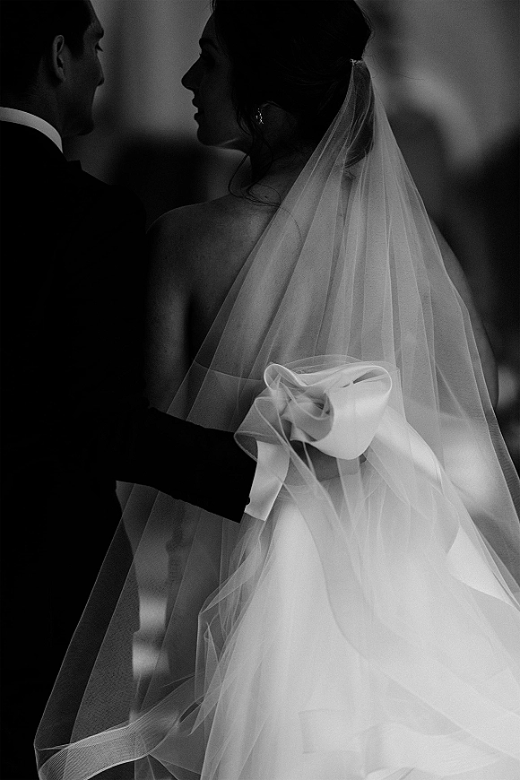 Couple portrait in a black and white wedding photo, bride and groom forehead to forehead, her bridal veil bow visible against a blurred indoor background