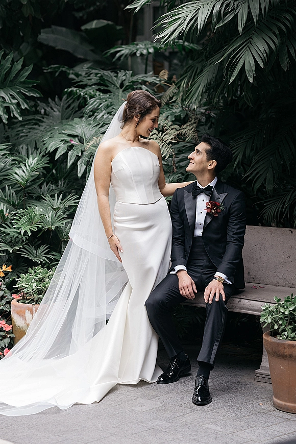 Couple portrait of bride in a strapless wedding dress with cathedral veil beside groom in tuxedo on a garden bench amid tropical greenery