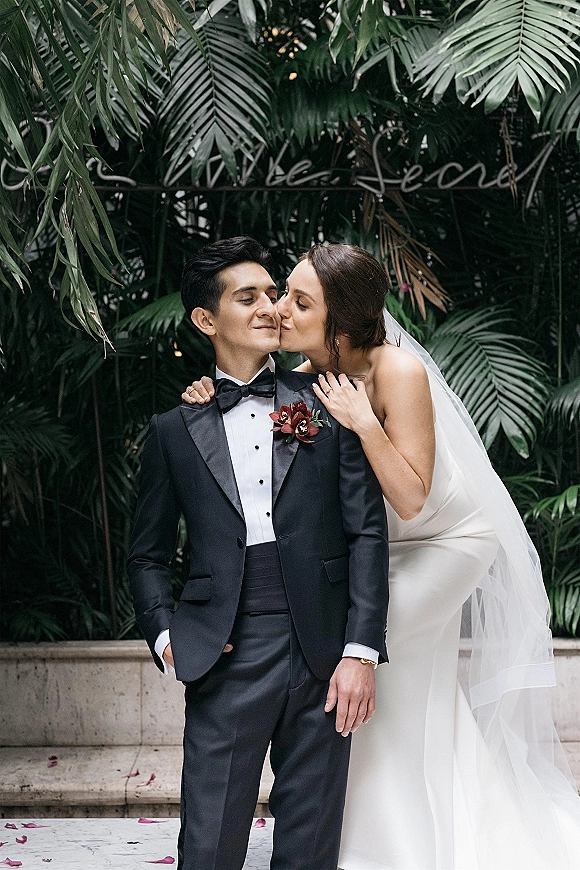 Wedding couple portrait with bride kissing groom’s cheek, her long veil draped as he wears a tuxedo and boutonniere against tropical greenery
