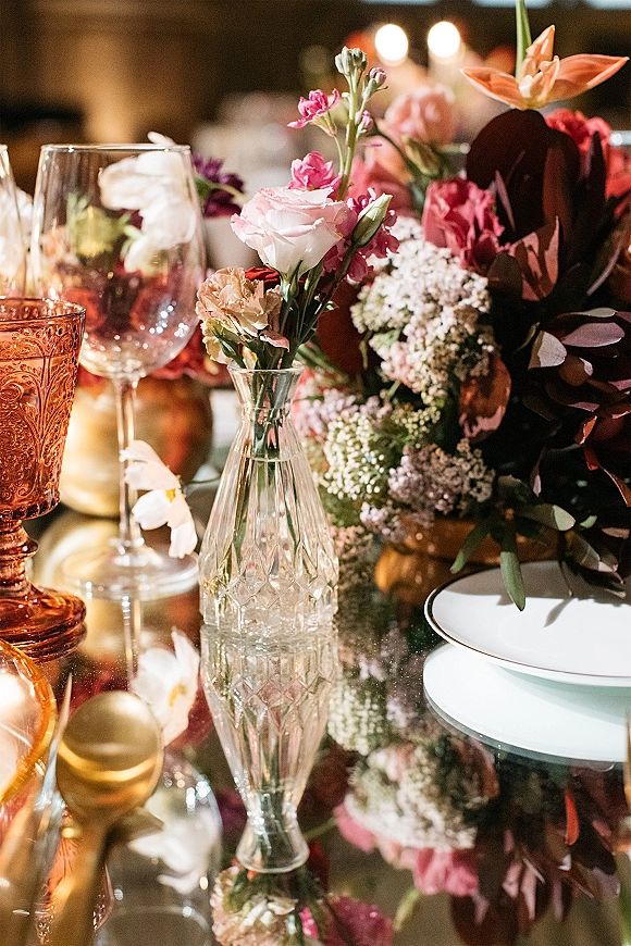 Wedding tablescape with wedding centerpiece ideas featuring mixed blooms in a clear bud vase, wine glasses and amber goblet on mirrored tabletop under bistro lights