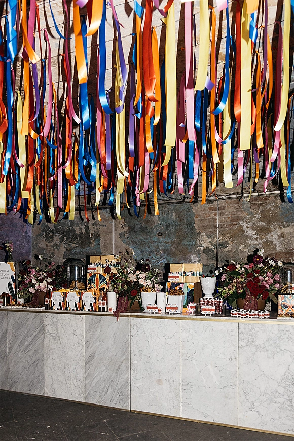 Wedding dessert table with a wedding candy buffet, ribbon streamers, floral arrangements, and favor jars on a marble counter against brick wall