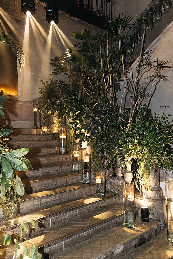 Wedding stairway decor with glass cylinder vases and pillar candles, greenery garland, and uplighting along a stone staircase and railing