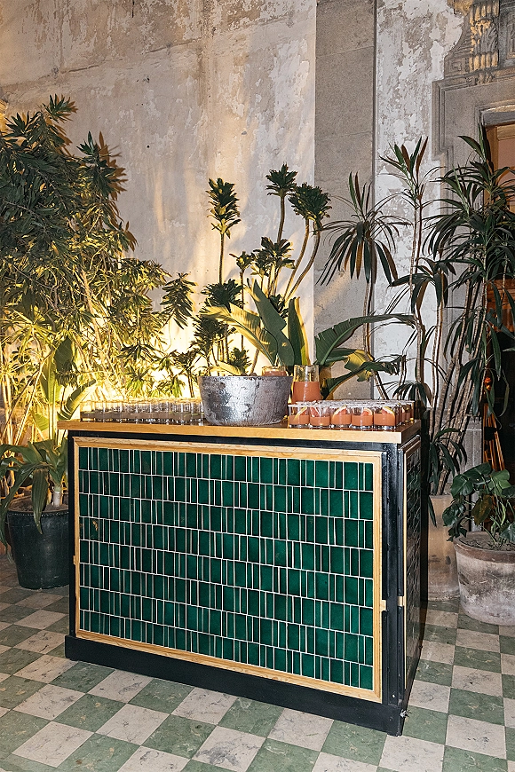Wedding bar setup with tiled bar front, cocktail glasses and orange drinks beside a metal ice bucket, framed by potted plants and uplighting indoors