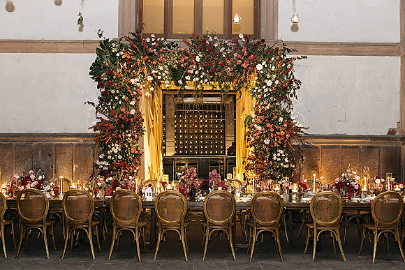Reception tablescape with head table decor, taper candles, glass vases, and lush florals beneath a draped floral arch in an industrial hall.