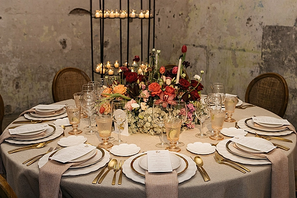 Reception tablescape with round wedding table setting, floral centerpiece and taper candles, gold flatware and goblets against plaster wall