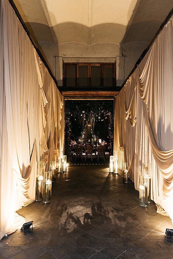 Wedding reception entrance with draped hallway wedding decor, white curtains, candle cylinders, and uplighting along a stone tile corridor under arches