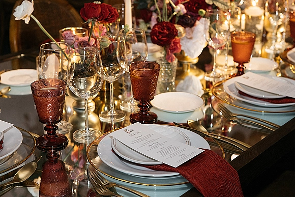 Reception tablescape with wedding place setting featuring gold-rim charger plates, red napkins, amber goblets, and candlelit floral centerpiece in dim lighting