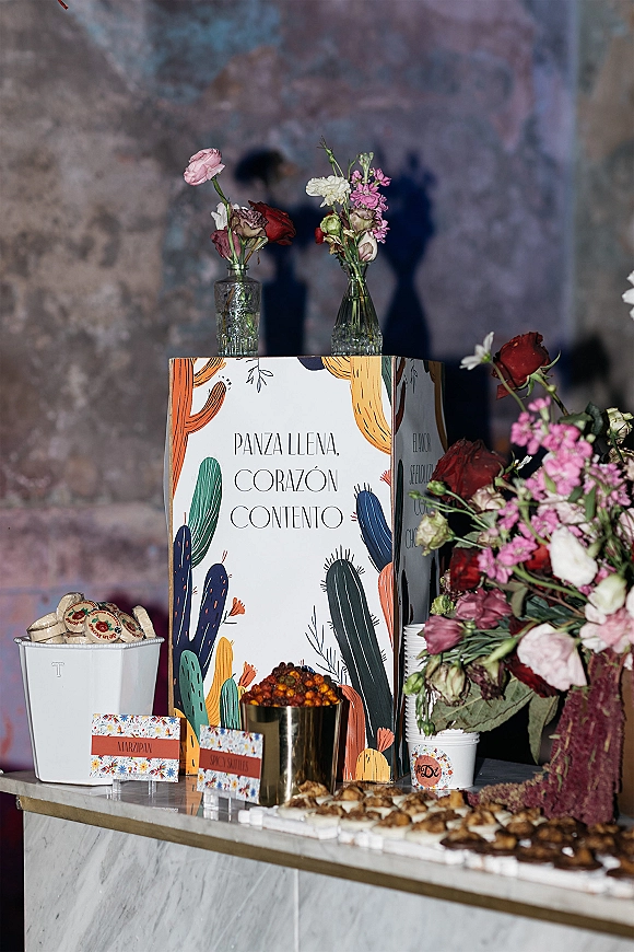 Wedding dessert table with wedding dessert display of marzipan cookies, place cards, bud vase florals on a marble counter by a stone wall