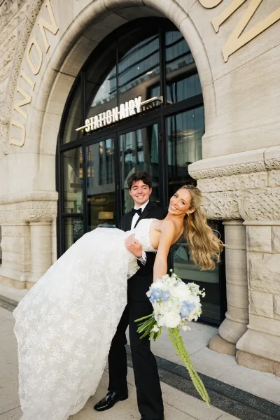 Couple portrait with the groom carrying the bride, her bouquet framed by a grand arched stone entrance and glass doors.