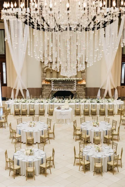 Reception decor with long head table styling and lush florals, set before a grand stone fireplace on a tile floor beneath a high ceiling.