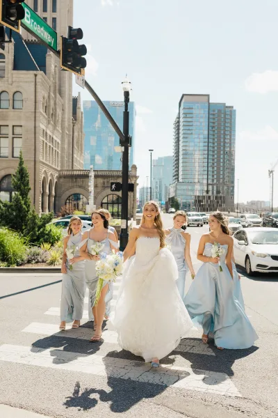 Bride and bridesmaids, a bridal party walking across a city crosswalk, laughing with bouquets as skyscrapers frame a nearby historic building.