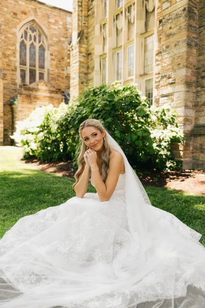 Bridal portrait of a bride in a sitting pose on a grass lawn, her veil draped softly, before a stone building with arched windows.