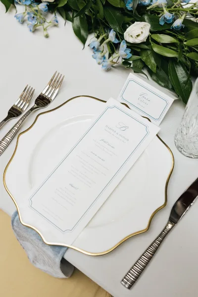 Wedding menu, a blue and white menu card resting on a charger plate, against a white tablecloth with green leaves and blue blossoms.