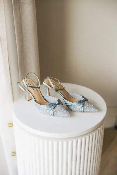 Bridal shoes, blue wedding heels with slingback straps, displayed on a white pedestal table against a beige wall and linen curtain.
