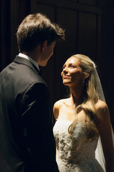 Couple portrait of a bride and groom facing each other, her long veil catching soft light against a warm wood panel wall.