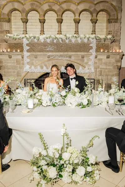 Couple portrait of newlyweds at a sweetheart table, smiling before a stone fireplace with arched niches and a candlelit mantel.