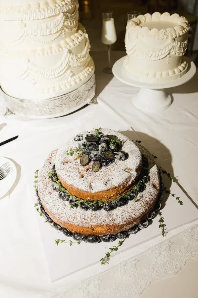 Wedding cake styled as a fruit topped wedding cake, crowned with grapes on a white tablecloth beside cake stands and champagne flutes.