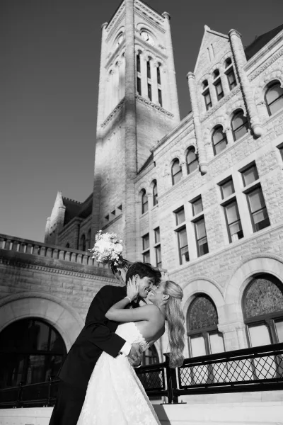 Couple portrait of the groom dipping the bride, her bridal bouquet lifted, framed by stone architecture and a clock tower with arched windows.