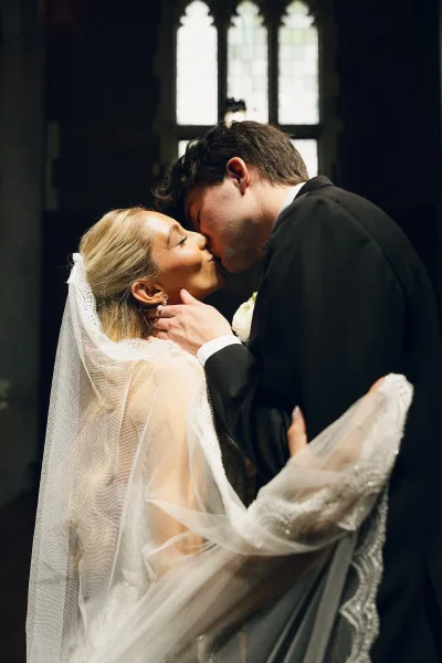 Wedding kiss portrait as the couple share their ceremony first kiss, with her bridal veil, framed by stained glass windows in a church interior.