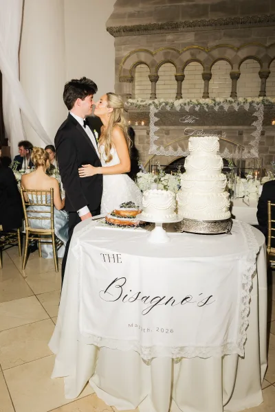 Wedding kiss portrait of a couple by the tiered buttercream wedding cake before a stone fireplace, guests softly blurred behind.