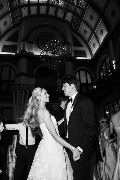 Bride and groom dance during their wedding first dance beneath a glowing chandelier in a ballroom interior crowned by an ornate ceiling.