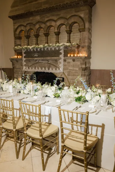 Reception tablescape styled as long banquet table decor, highlighted by a lush floral garland, set before a stone fireplace with an arched mantel.