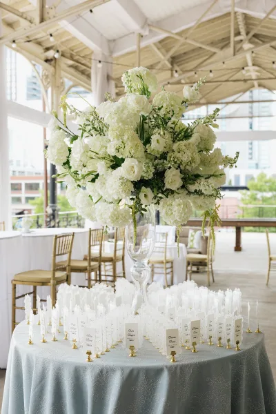 Escort card display as a seating chart alternative, anchored by a tall glass vase in an open-air pavilion with string lights and city views.