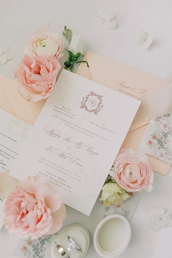 Invitation suite wedding invitation flat lay with blush envelopes, calligraphy crest, floral liner, ring box and rings on fabric with petals