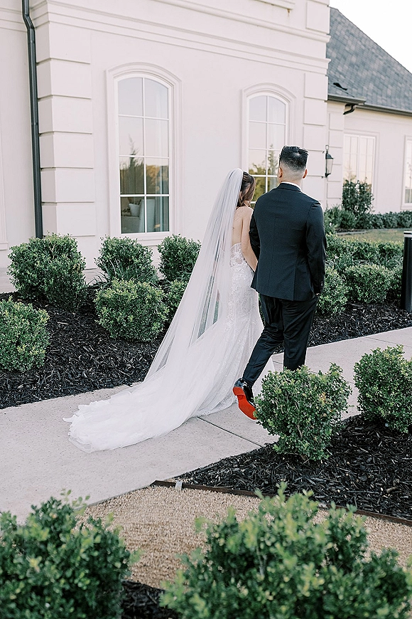 Couple portrait of bride and groom walking away, her lace gown and long veil trailing behind on a walkway by a white building