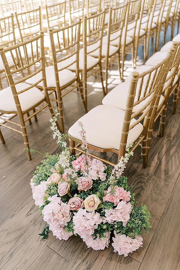 Ceremony seating with gold chiavari chairs and ivory cushions, lined along a sunlit wood floor aisle with rose florals.