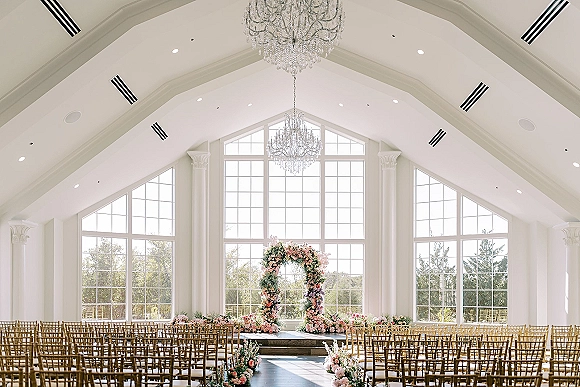 Ceremony setup for an indoor wedding ceremony with a floral arch of blush blooms and greenery, gold chiavari chairs under chandeliers