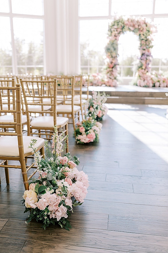 Ceremony setup with blush pink floral arch and gold chiavari chairs, aisle flowers lining a windowed indoor space in natural light