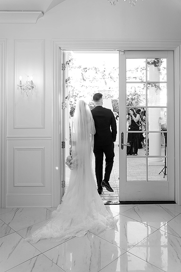 Wedding processional as bride and groom walk away through glass doors, bridal veil trailing over gown train in an elegant marble hallway
