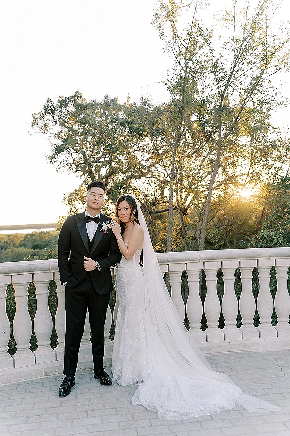 Couple portrait of bride in a wedding dress with long veil and groom in tuxedo on a stone terrace at sunset with trees and water beyond