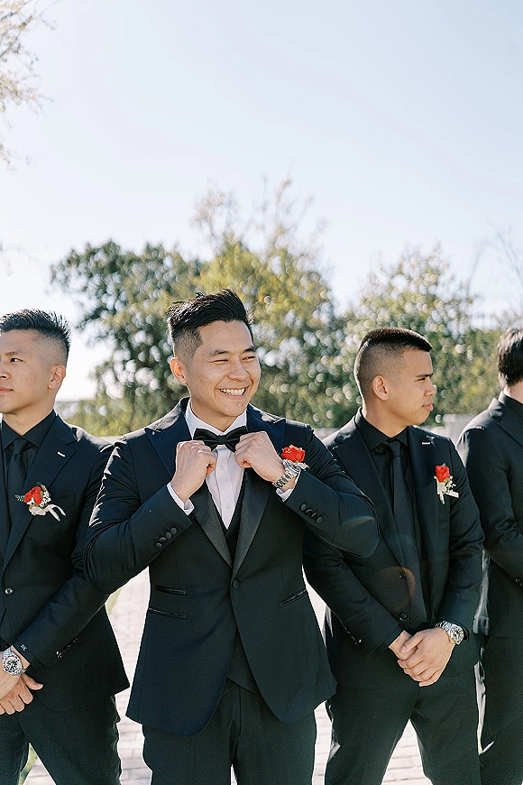 Groomsmen portrait of the groom with groomsmen in black tuxedos and bow ties, with red boutonnieres, outdoors under sunny trees and blue sky