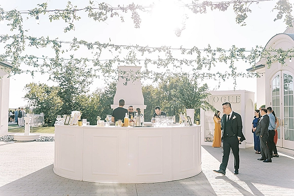 Wedding bar setup with round wedding bar, glassware, liquor bottles, and framed signs under string lights on a sunny patio lawn