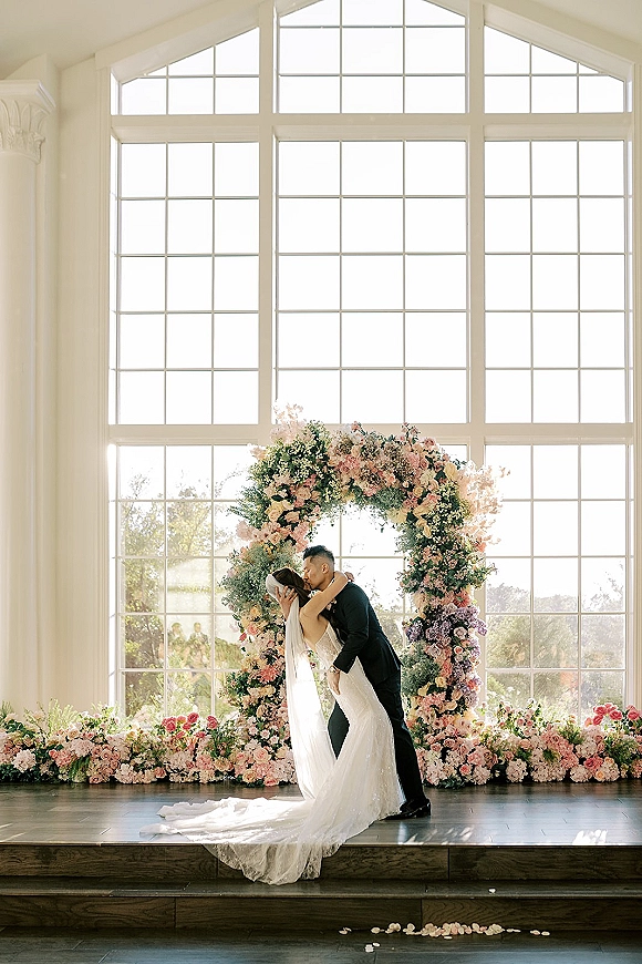 Wedding kiss portrait of bride and groom kiss under a pastel floral arch with pink roses, in natural light before tall arched windows