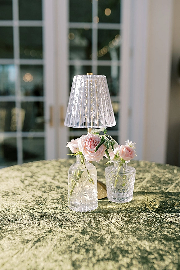 Wedding table centerpiece with a small brass lamp, blush pink roses in glass bud vases on a velvet tablecloth by window panes and warm bokeh lights