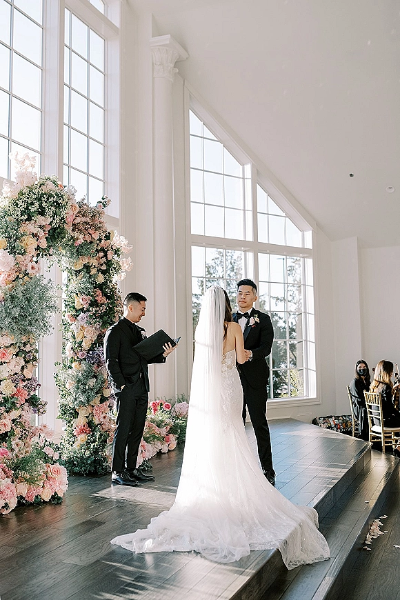 Wedding vows as bride in lace dress and veil faces groom in black tux beneath pastel floral arch in a sunlit chapel with tall windows