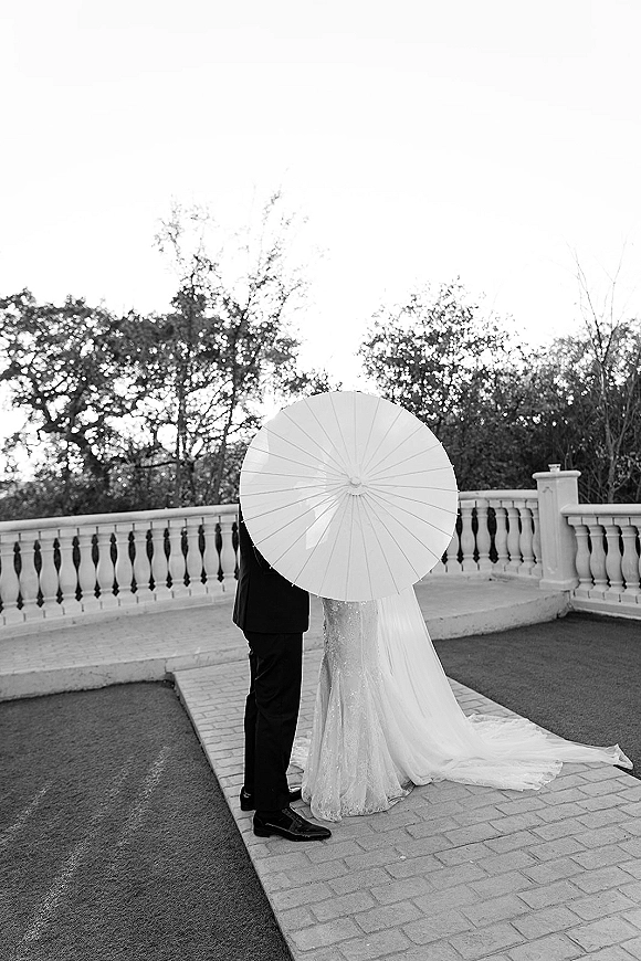 Wedding couple portrait of bride and groom under a white parasol, her veil and gown train flowing on a stone terrace by balustrade.